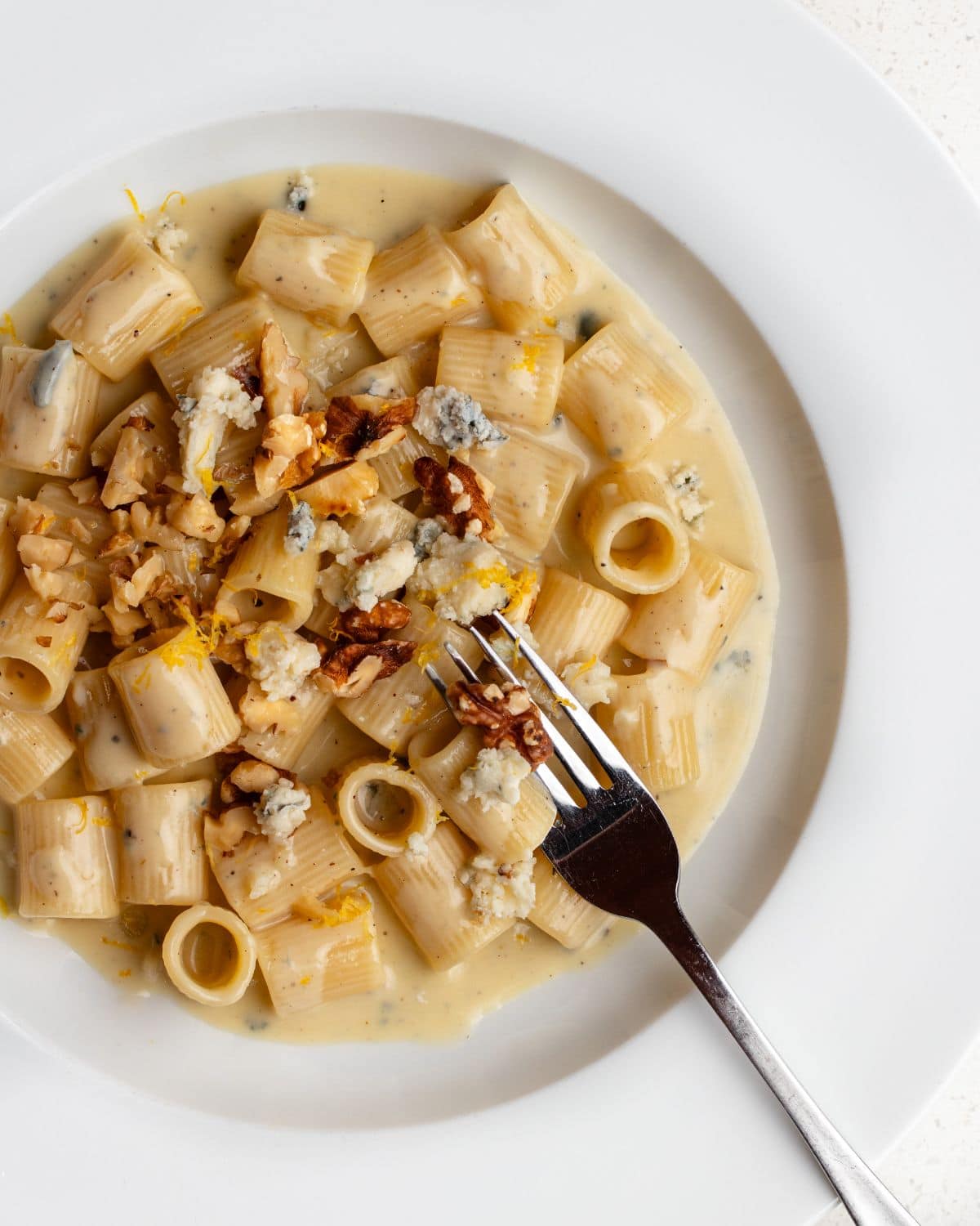 White plate with rigatoni pasta in a creamy gorgonzola sauce. Walnuts and lemon zest on top.