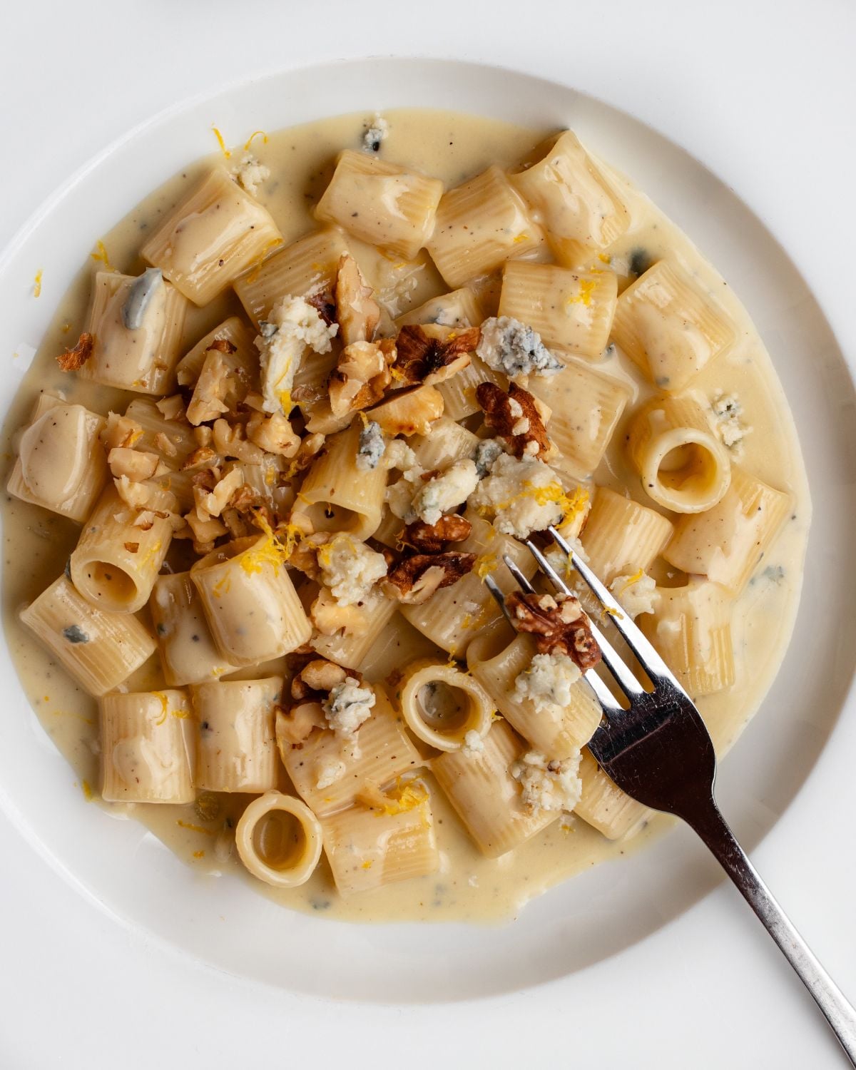 White plate with rigatoni pasta in a creamy gorgonzola sauce. Walnuts and lemon zest on top.