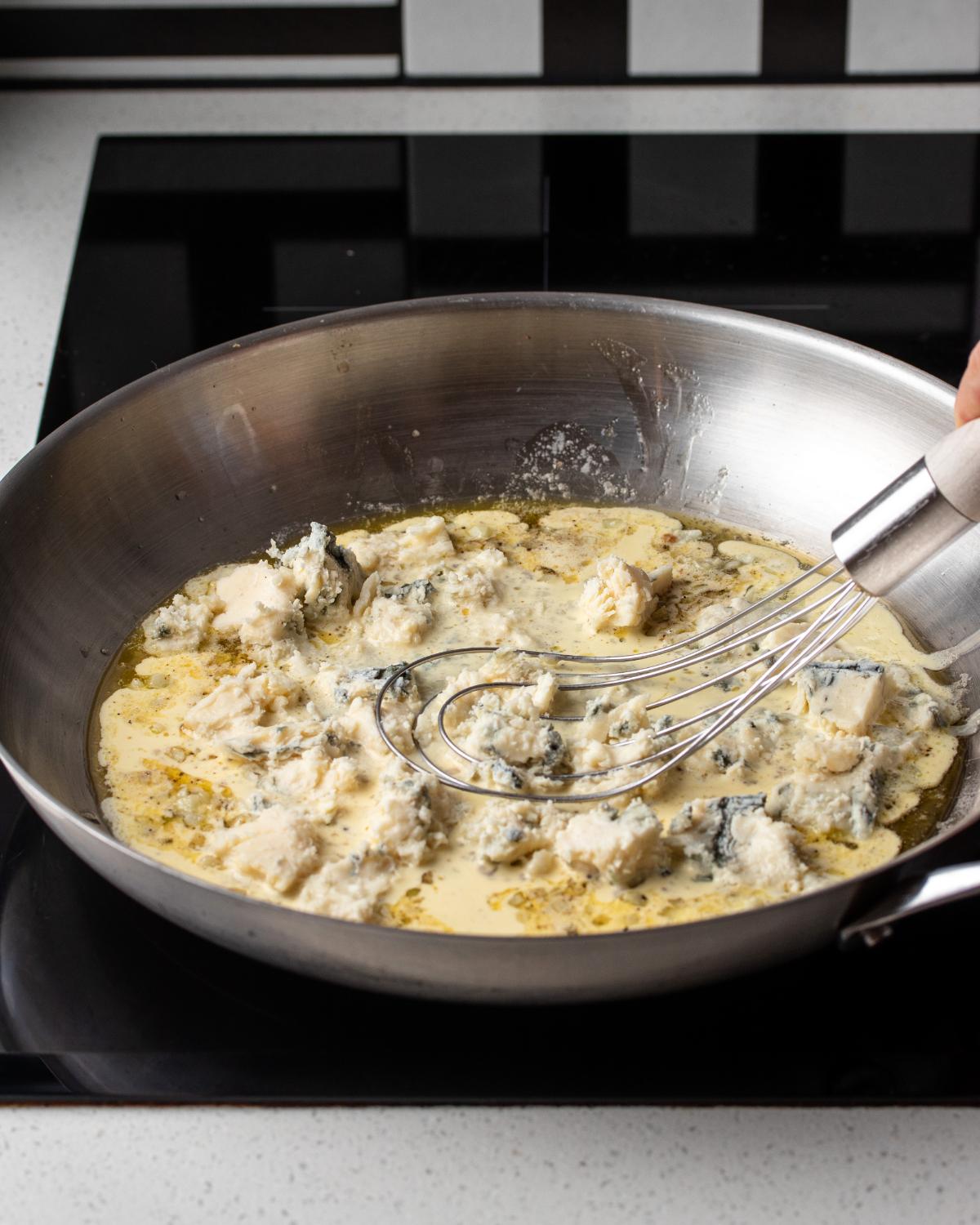 Gorgonzola cheese melting into heavy cream in a skillet, whisk resting.