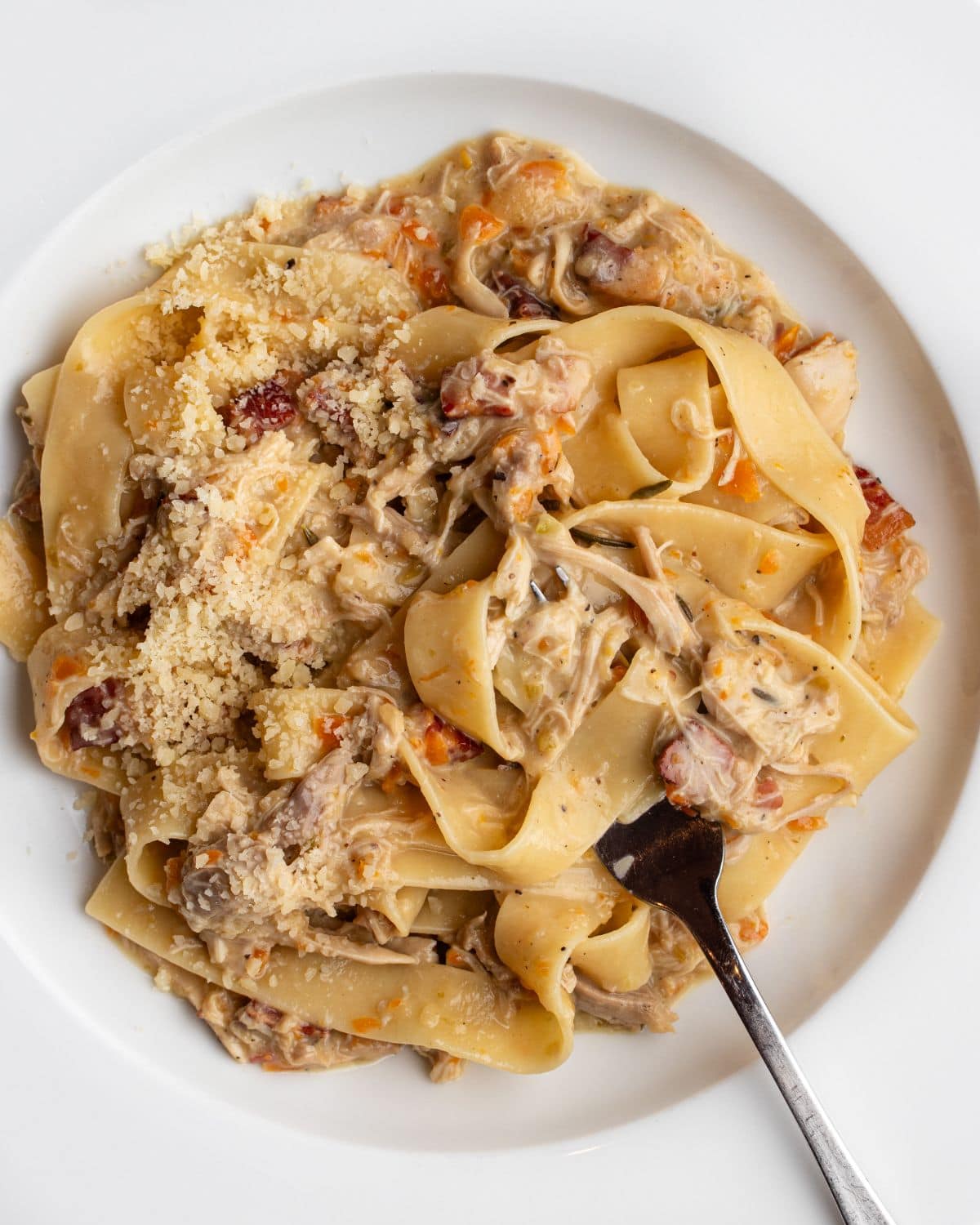 White plate with pappardelle noodles in a chicken ragu. Fork resting into pasta.