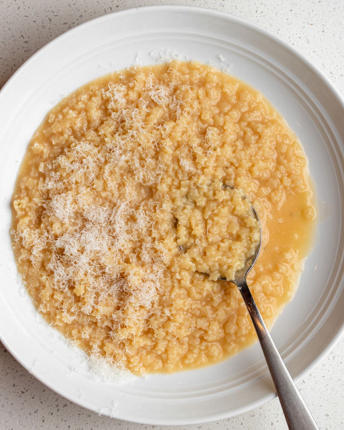 A white bowl with pastina served and a spoon resting in. Grated parmesan on top.